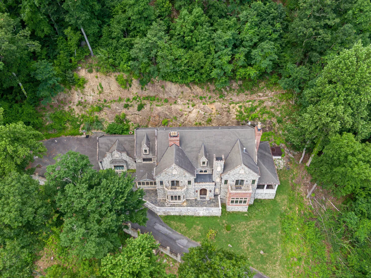A wide exterior view of the stone mansion surrounded by mature trees, with a stately driveway leading to the entrance. The enigmatic mood, sunset illumination, and natural surroundings hint at the estate’s grandeur, longstanding presence, and connection to the Blue Ridge landscape.