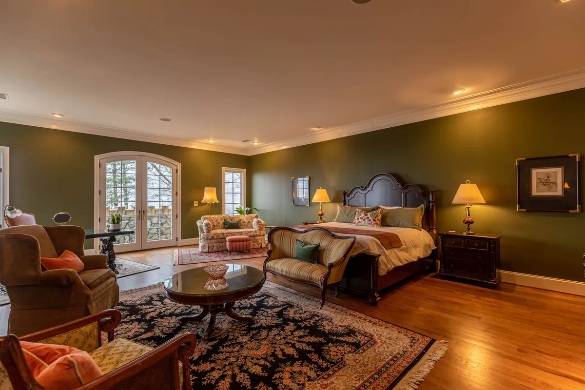 A luxurious bedroom featuring an ornate headboard, French doors opening to a private balcony, decorative lamps, a round wooden coffee table, and a richly patterned area rug. The room exudes warmth and elegance, with plush bedding and antique furnishings.