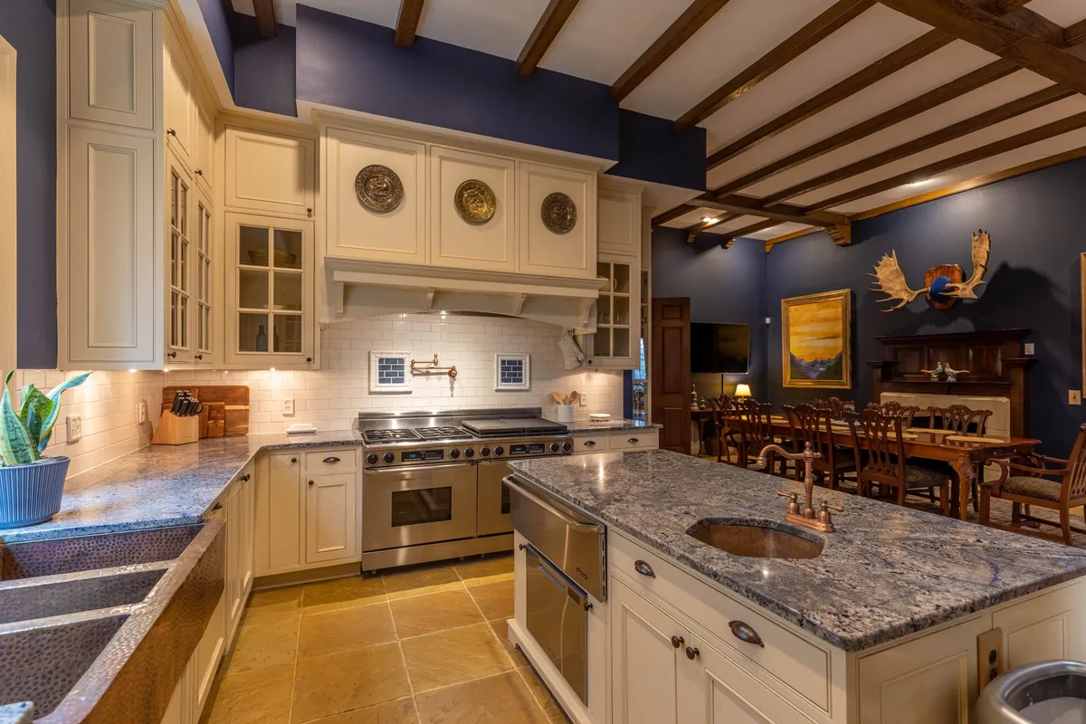 The image features a warm and inviting kitchen with a granite countertop, stainless steel appliances, and a dining area visible in the background. Wooden beams overhead add rustic elegance, and the space is clearly designed for both cooking and seamless entertaining.