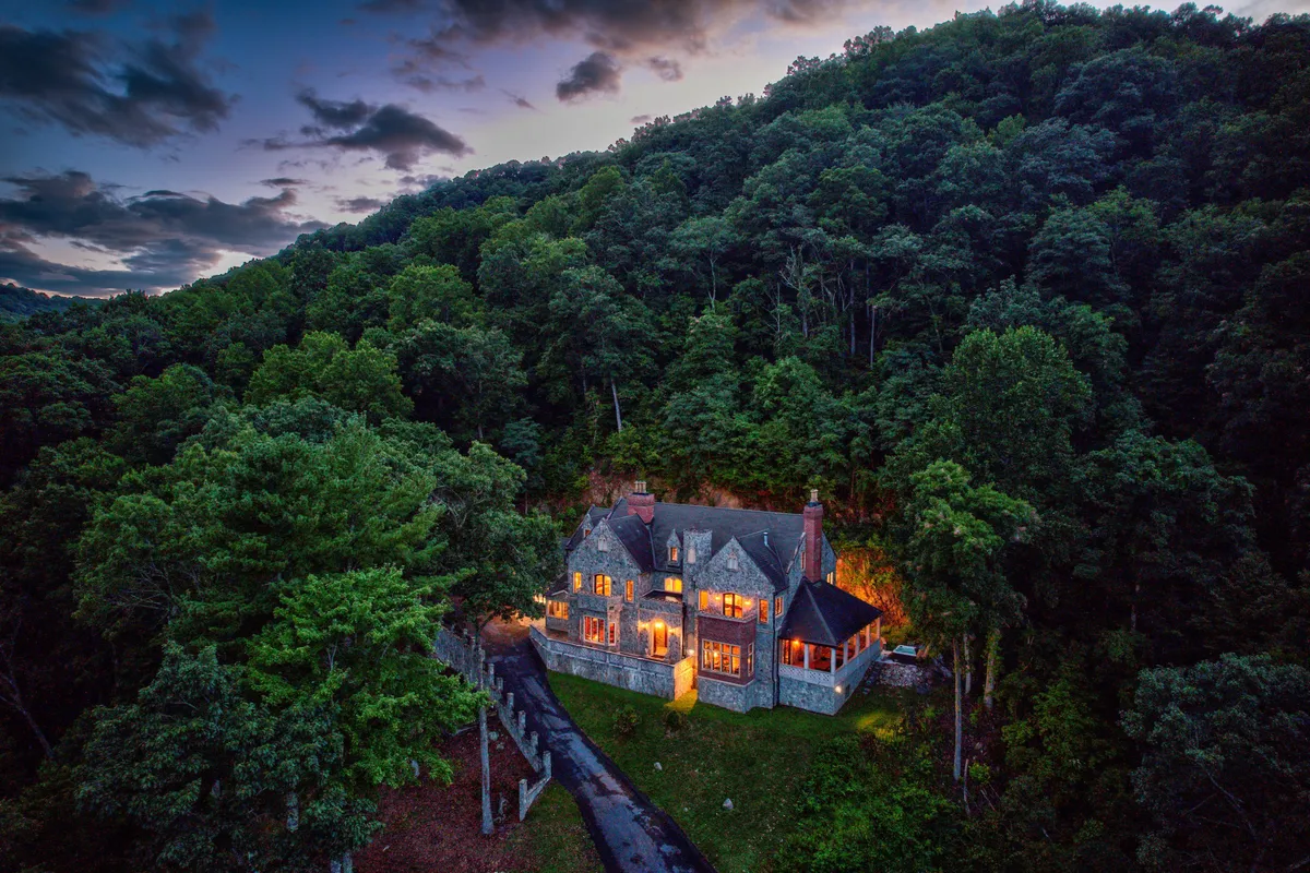 A majestic view of the mansion with its glowing windows set amidst a lush forest. The exterior is warmly lit, suggesting a lively evening inside, while the surrounding woods invite guests for nature walks and outdoor exploration.