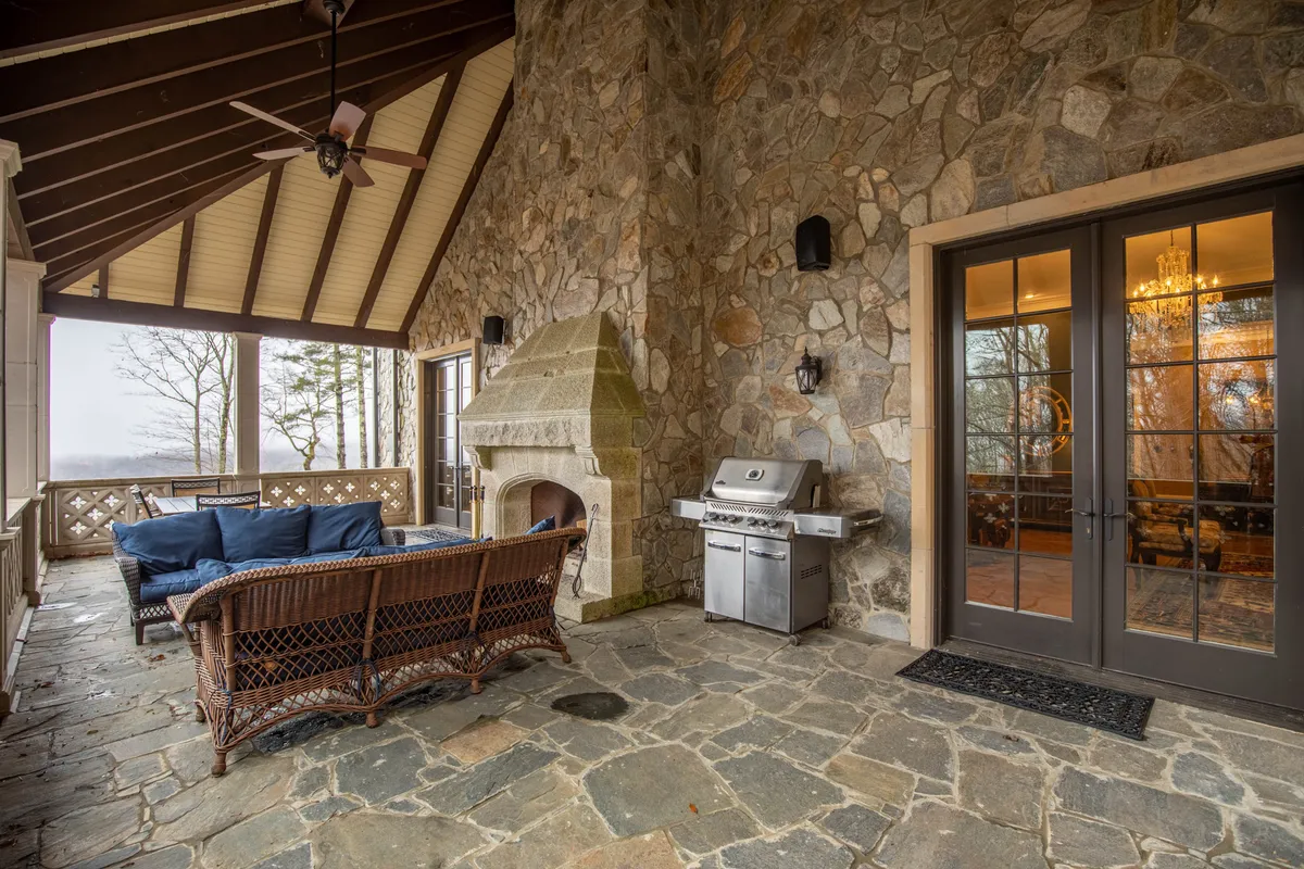A covered verandah with comfortable outdoor seating, a classic stone fireplace reminiscent of old-world European charm, an outdoor grill, and double doors opening to the interior, all framed by nature.