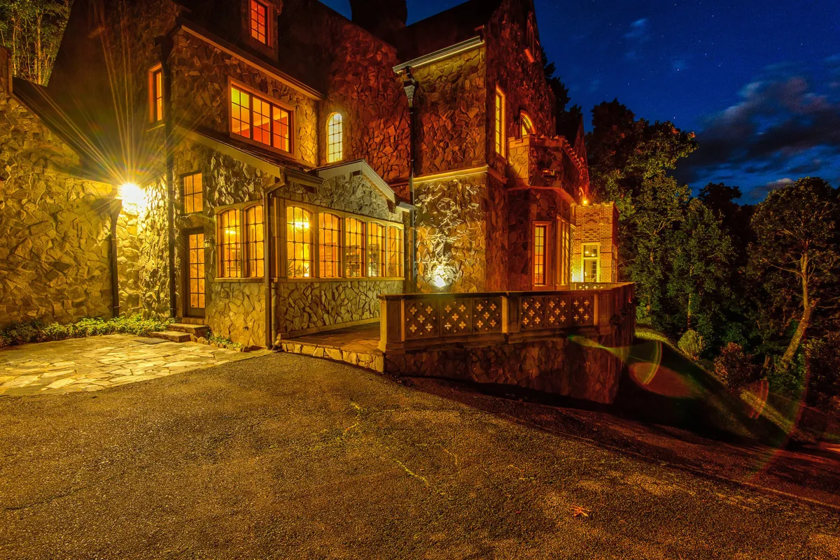 An enchanting exterior evening view of the castle, its windows warmly aglow against the stone facade and balcony railing. The scene suggests cozy indoor moments while referencing the outdoor environment.