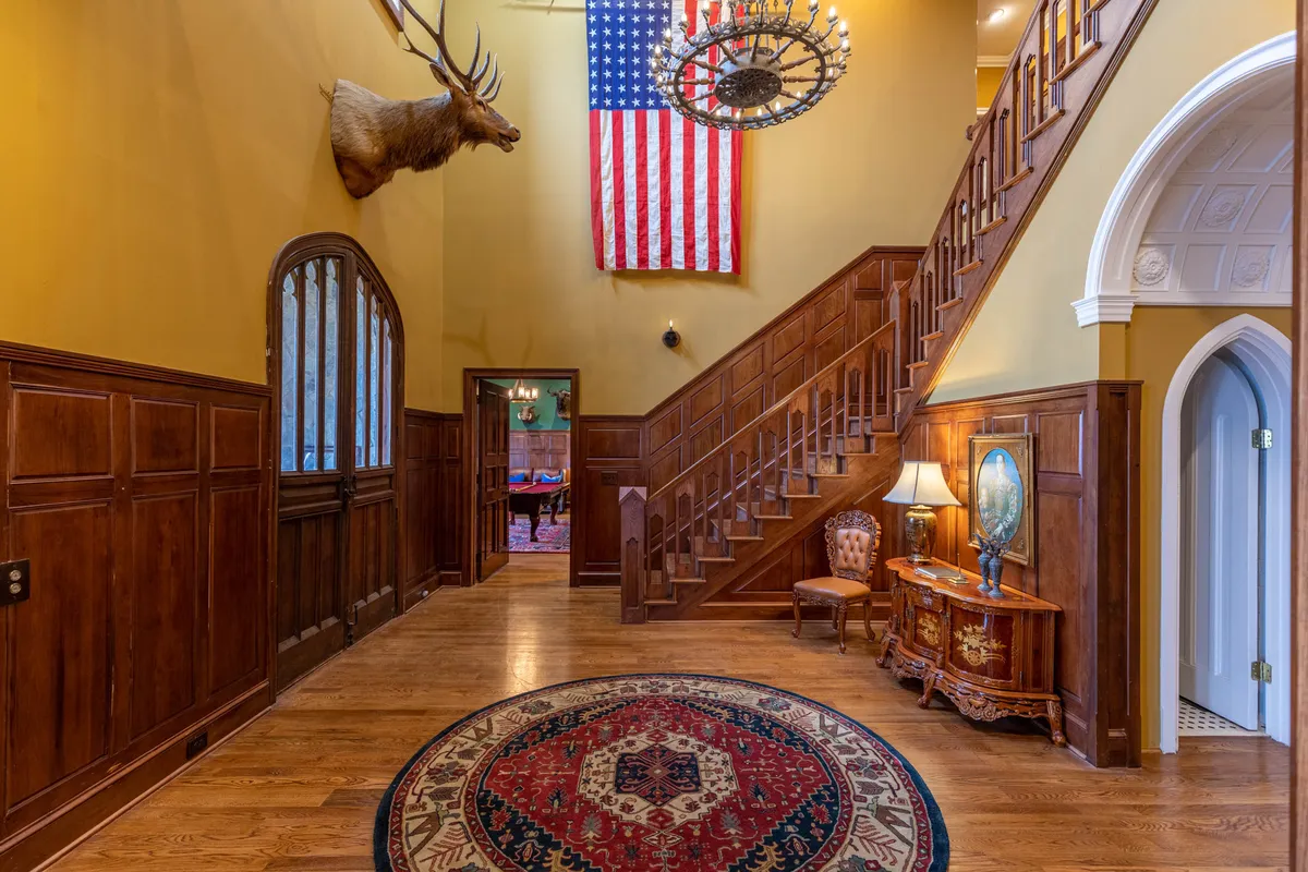 A grand foyer featuring an ornate chandelier, a mounted moose head above the entrance, an elegant staircase, and a decorative rug, all combining to create an opulent and welcoming first impression.