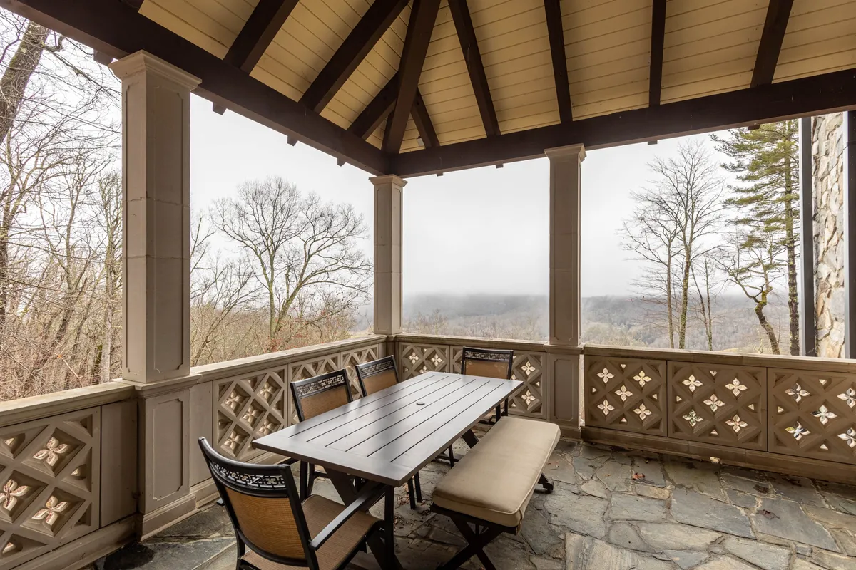 An inviting outdoor dining area with a wooden table, slate flooring, ornate railing, and a sweeping view of trees and distant hills. The setup suggests shared meals, morning coffee, or evening gatherings under the sky.
