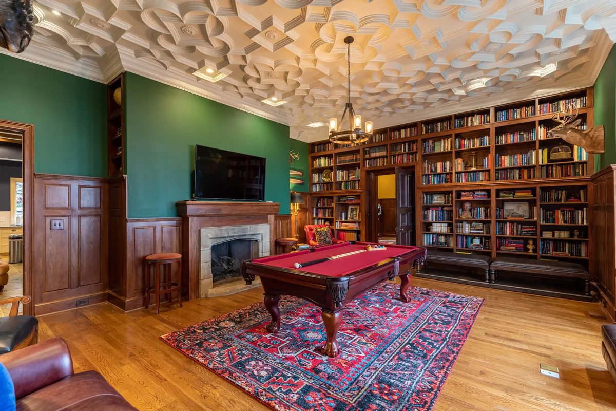 A luxurious library space featuring a pool table at the center, framed by elegant bookshelves filled with volumes, a grand chandelier overhead, and a warm fireplace. The rich wood tones and plush seating create an inviting setting for reading, relaxing, or enjoying a classic game of billiards.