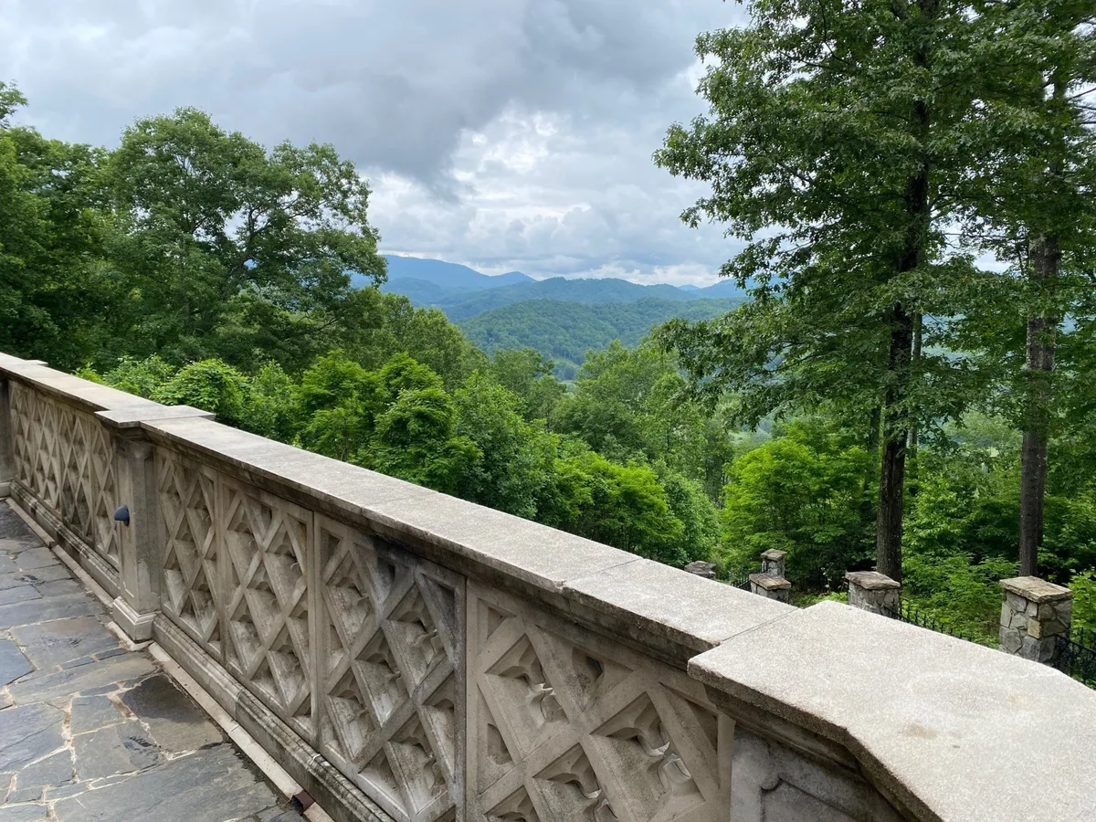 This image features an ornate stone railing in the foreground overlooking lush green trees and a mountain range in the distance. The tranquil setting is imbued with the golden hues of sunset, evoking a sense of peace and inviting outdoor enjoyment.