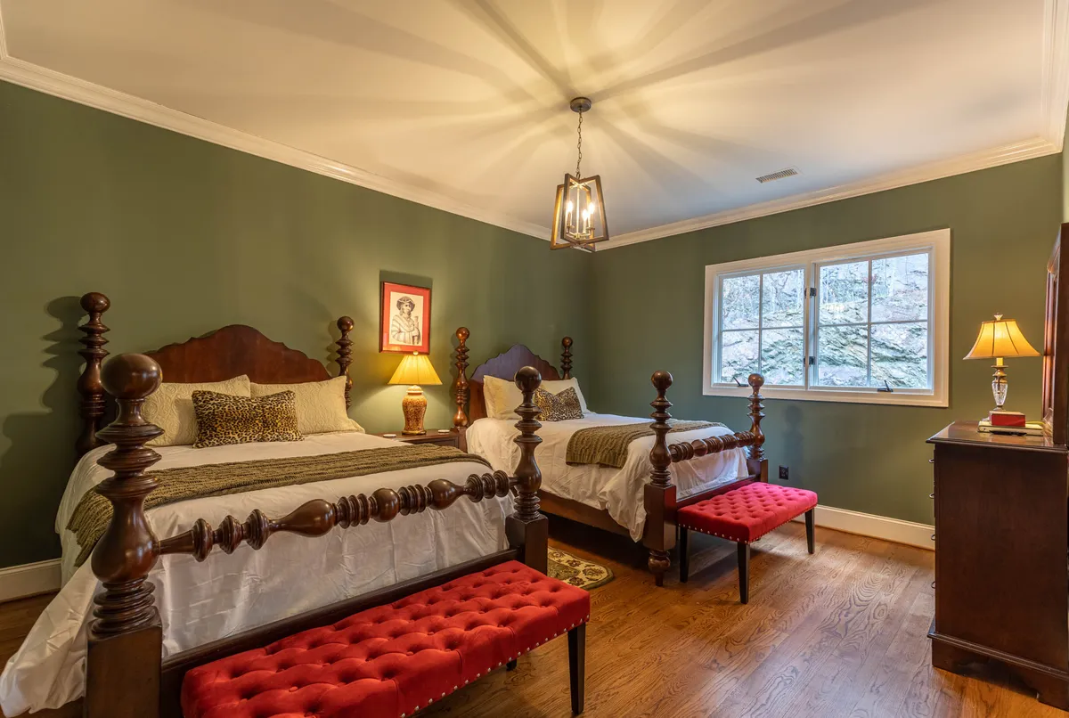 An elegant bedroom with two ornate wooden beds, red tufted benches, warm table lamps, and framed artwork. The space is both cozy and grand, ideal for family or group stays, with natural light streaming in.
